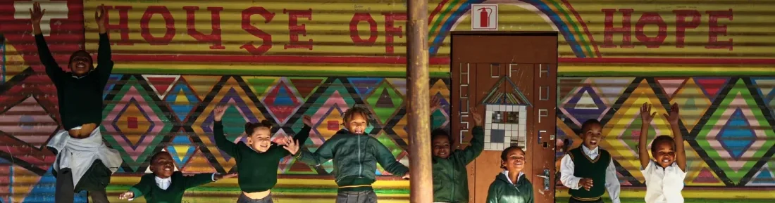 Children jumping in front of the painted House of Hope wall, arms in the air