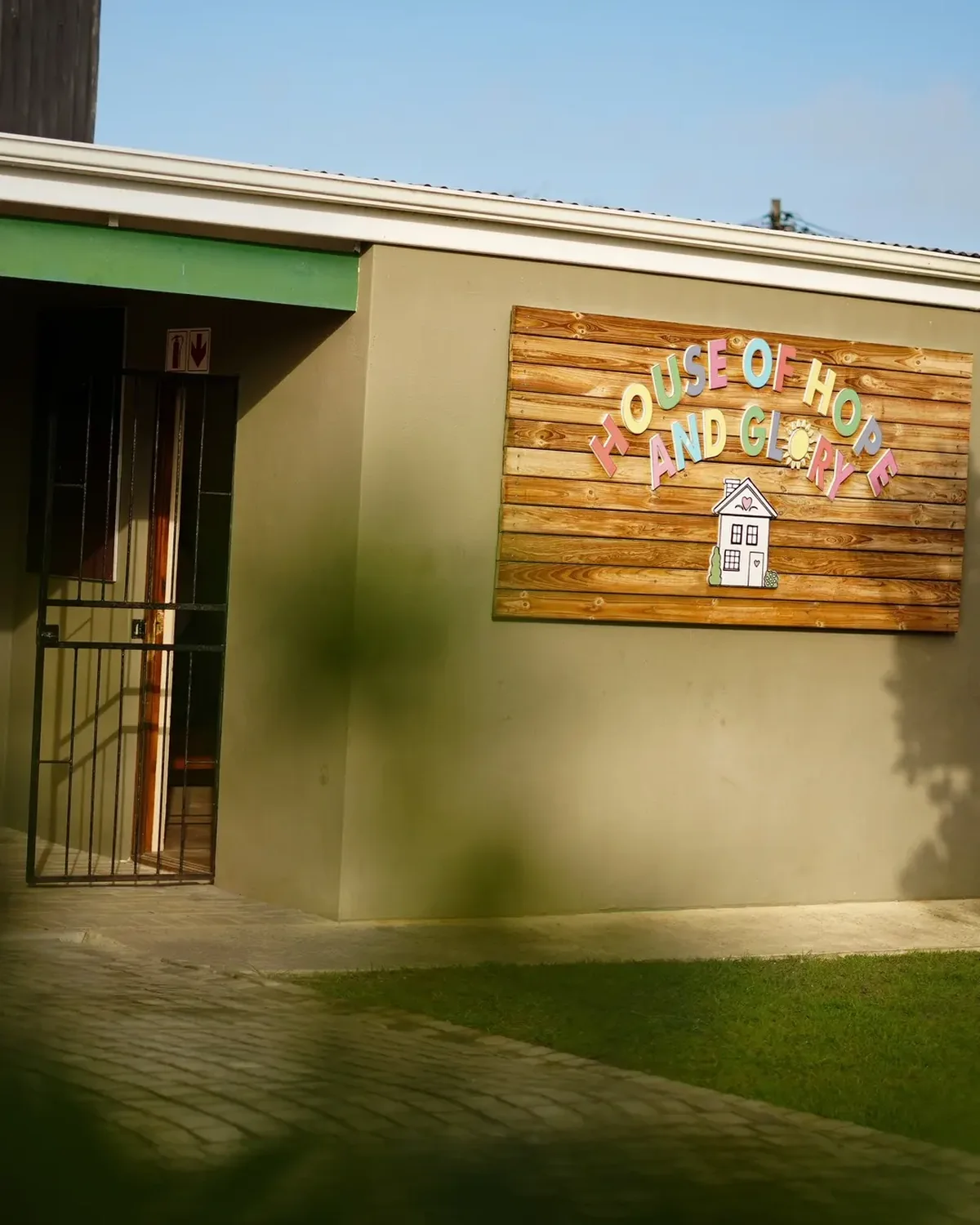 The House of Hope and Glory sign on the outside wall, hand-painted letters on wood above a small drawing of a house, with the gate standing open