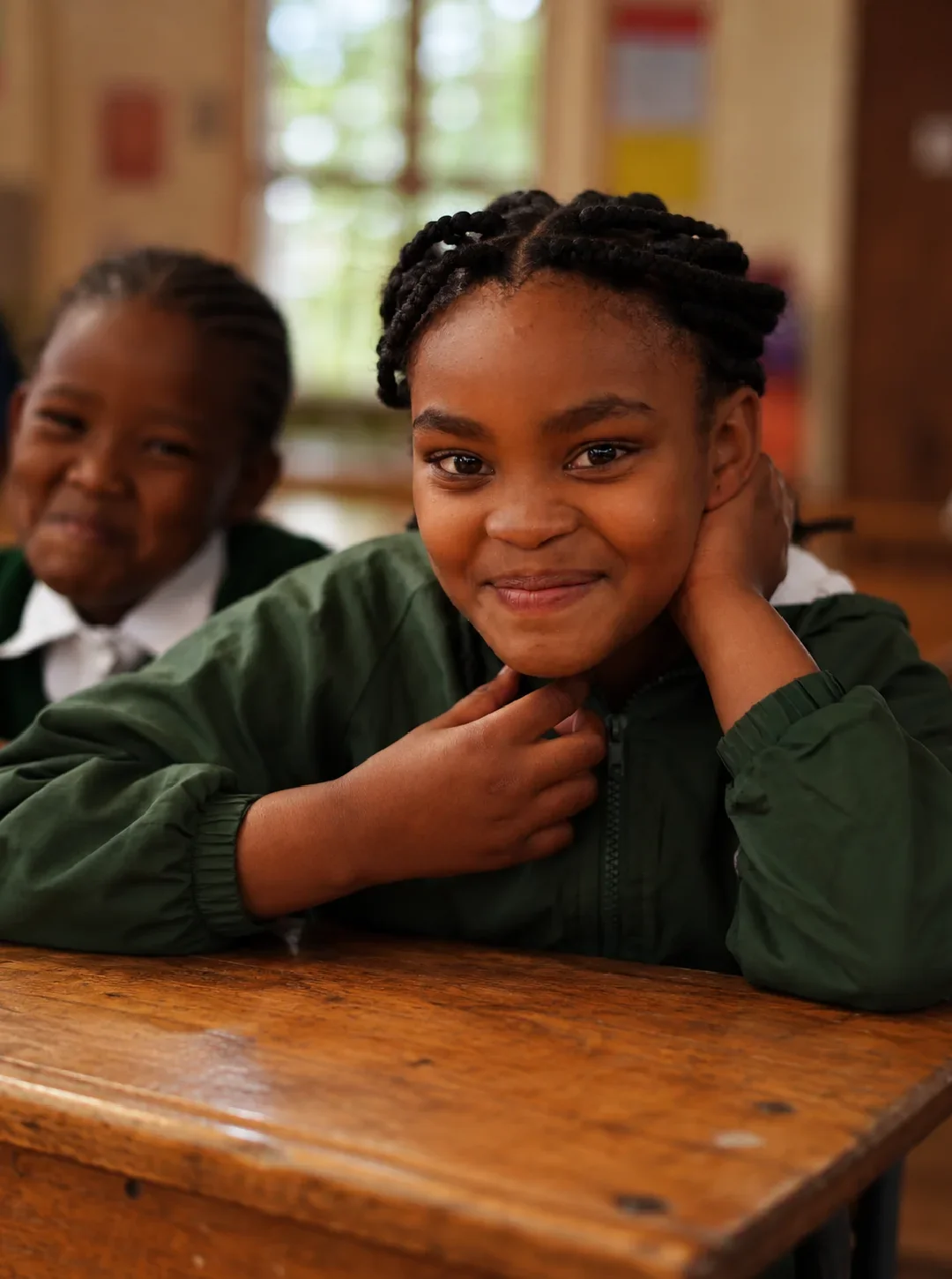 A girl resting her chin on her hands at a classroom desk, smiling at the camera, a friend beside her