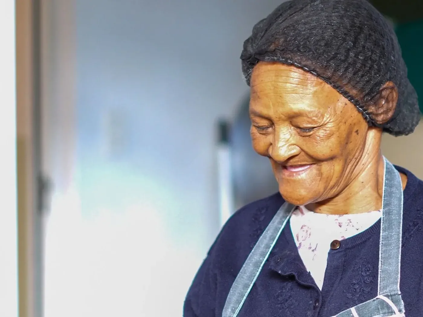 A cook in an apron and headscarf standing in the kitchen doorway, looking down and smiling