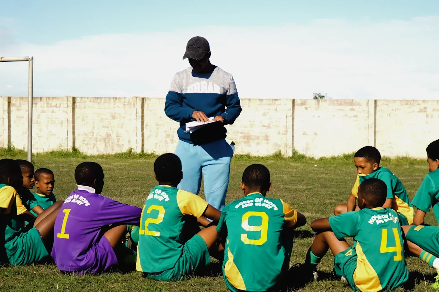 A coach with a clipboard talking to the team sitting on the grass in House of Hope and Glory kit